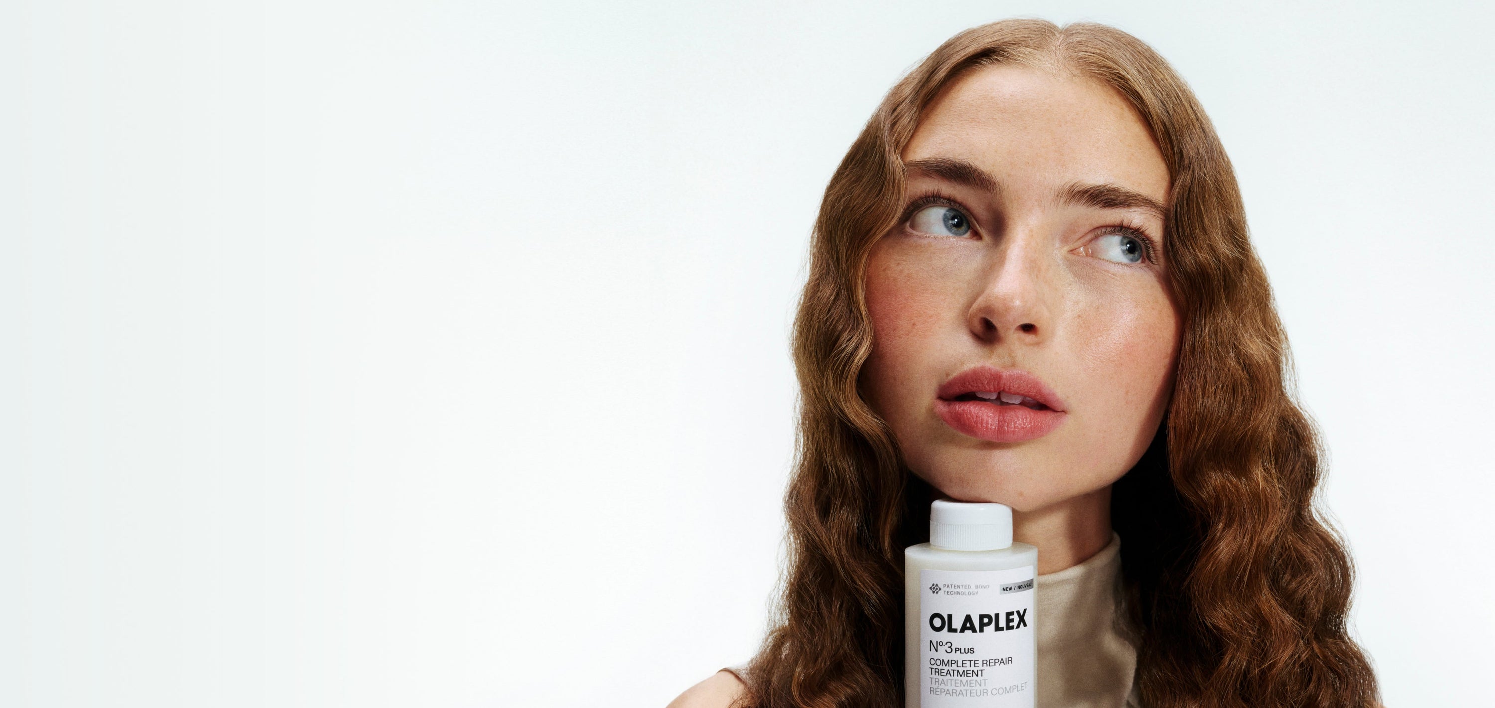 A woman with red hair holding the a bottle of OLAPLEX No.3PLUS under her chin