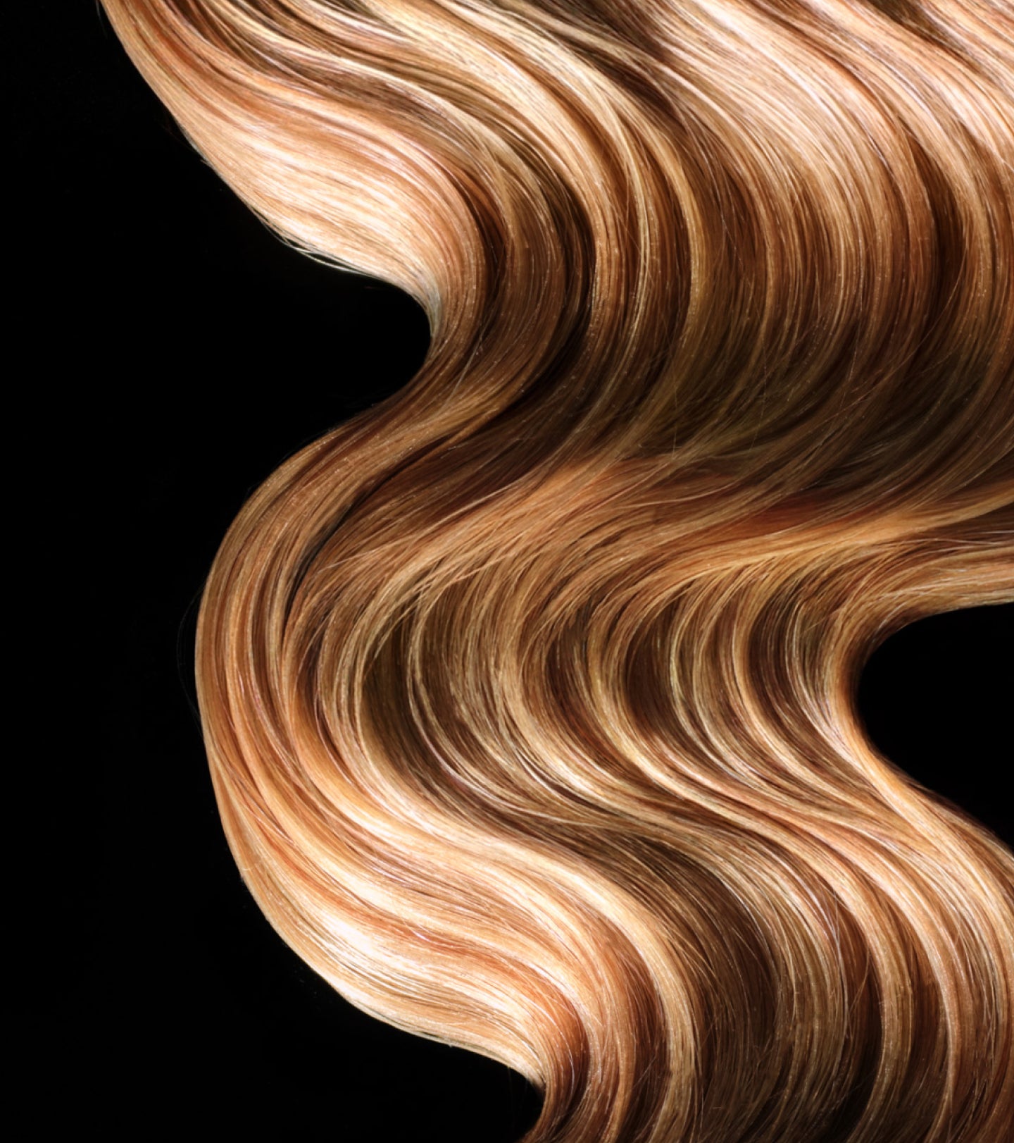 A close-up of flowing, wavy hair in a warm honey-blonde shade with lighter golden highlights, set against a solid black background.